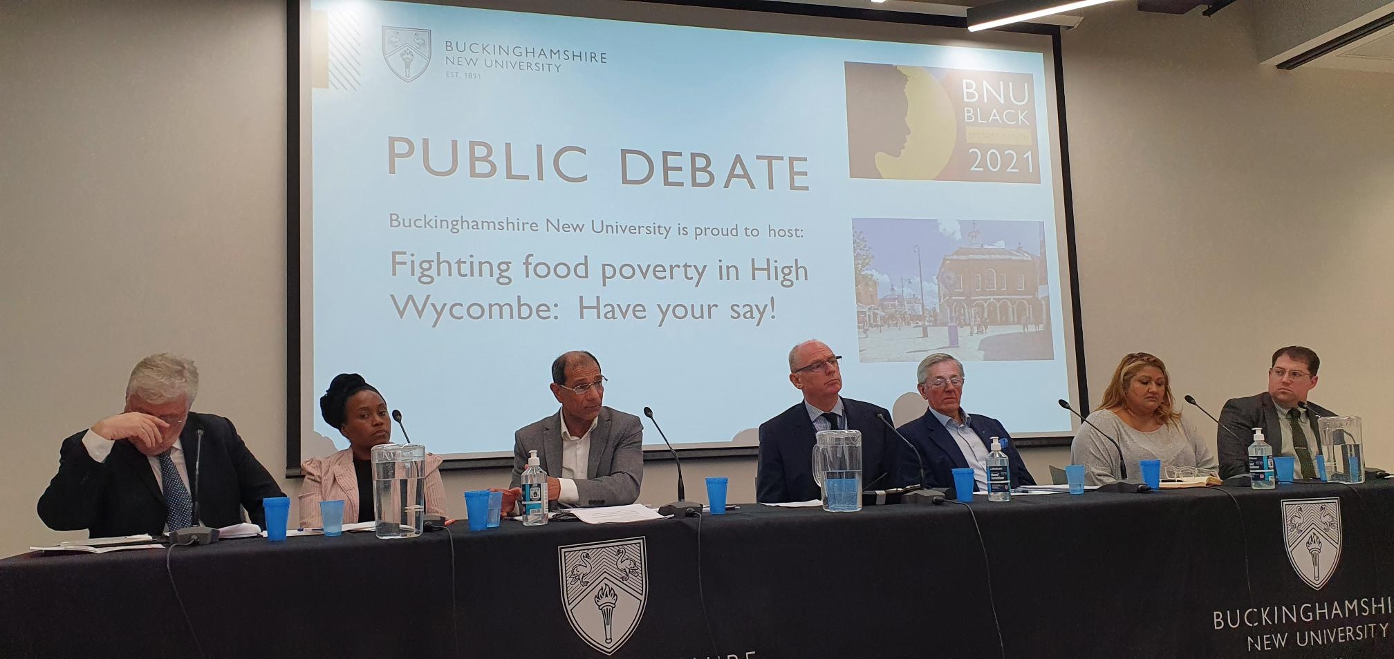 7 panellists sit at table at public debate